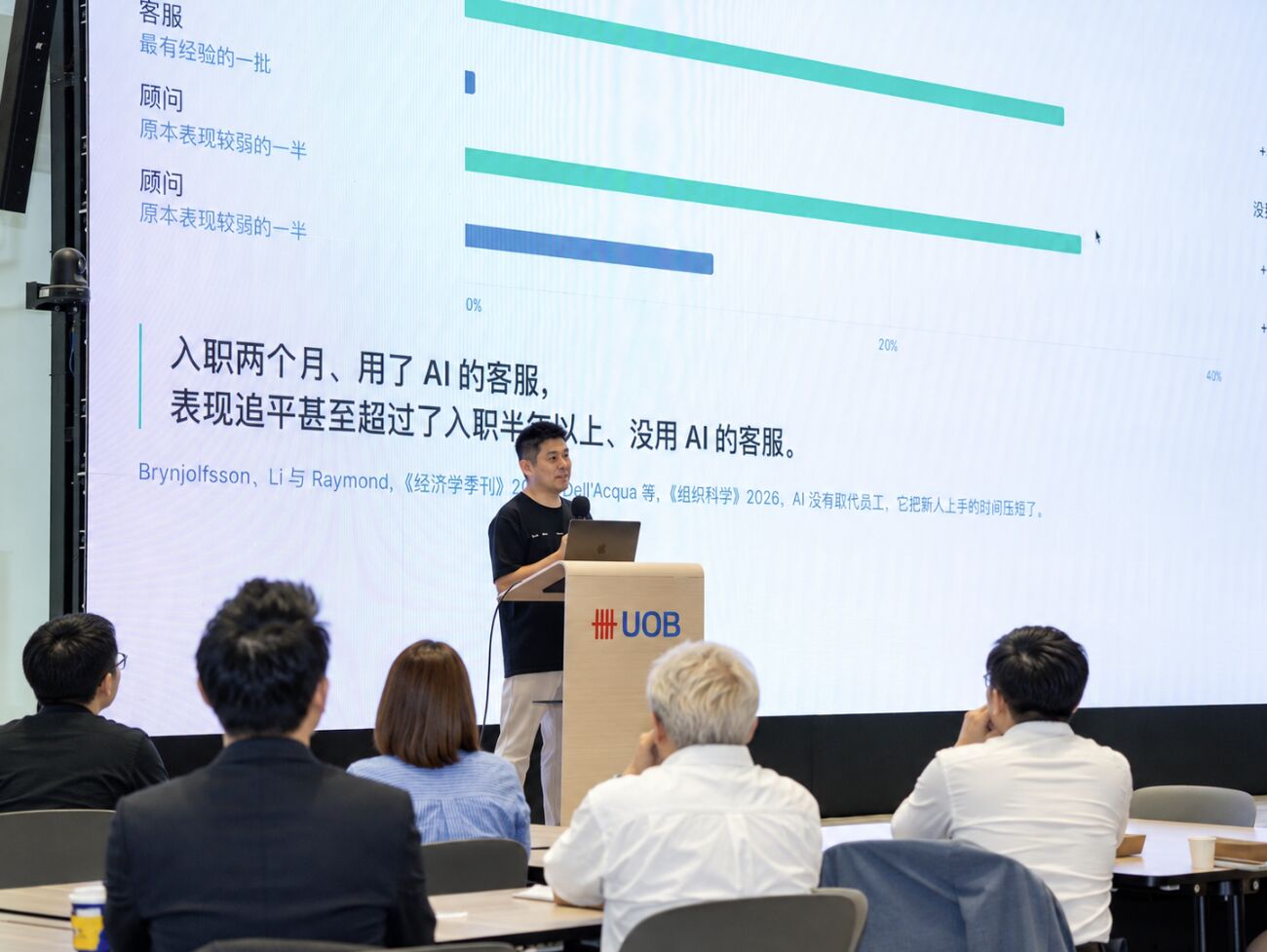 讲者在 UOB 讲台前演讲，身后的幻灯片写着：入职两个月、用了 AI 的客服，表现追平甚至超过了入职半年以上、没用 AI 的客服。